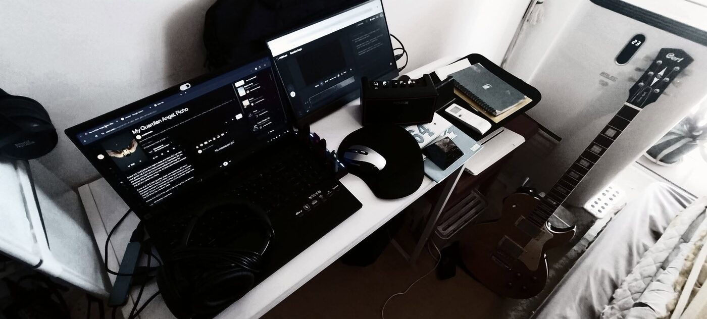 Paddy's bedroom music setup: a laptop showing the track My Guardian Angel by Paddy Picho, a second monitor, headphones, a small NUX amplifier, notebooks, and a Cort CR200 electric guitar leaning against a portable air conditioner beside the bed